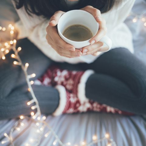žena sediaca doma, prekrížené nohy na posteli, držiaca šálku kávy, vianočná dekorácia