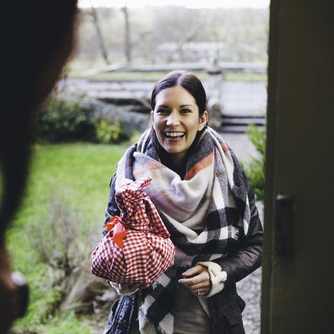ženska se nasmehne, ko stoji na vratih in obišče prijateljico, zavito je v topla oblačila in drži darilo, zavito v rdeče -belo krpo